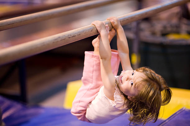 "Young girl, aged 2, proving it is never to early to start having fun and practicing good physical fitness. She is pulling herself up on uneven bars. Blue and yellow mats defocused in the background. Shallow DOF provides good subject isolation, focus on her eyes."